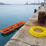 reddingsberrie-water-nooduitrusting-haven-redding.jpg Oranje SKED reddingsbrancard drijvend op kalm blauw water naast betonnen kade met reddingstouw in industriële haven