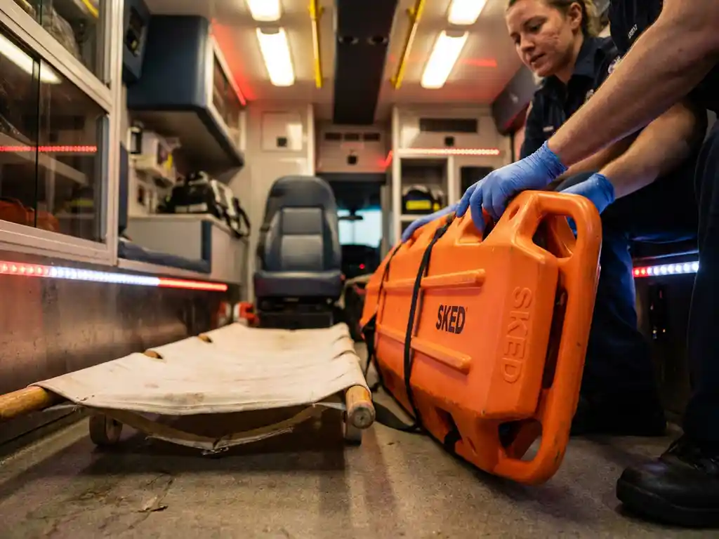 Oranje SKED evacuatiebrancards naast traditionele witte brancard in ambulance, hulpverlener past moderne reddingsuitrusting aan